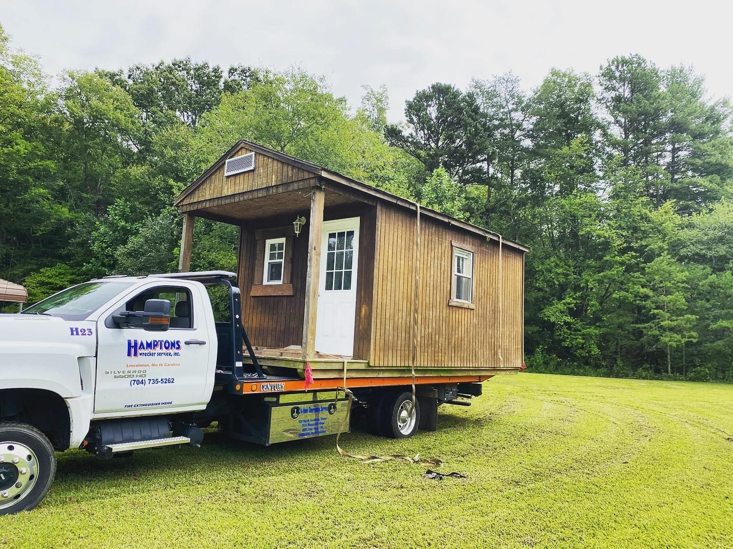 A small, rustic cabin being transported on a flatbed tow truck. The truck is in a grassy area with trees in the background.
