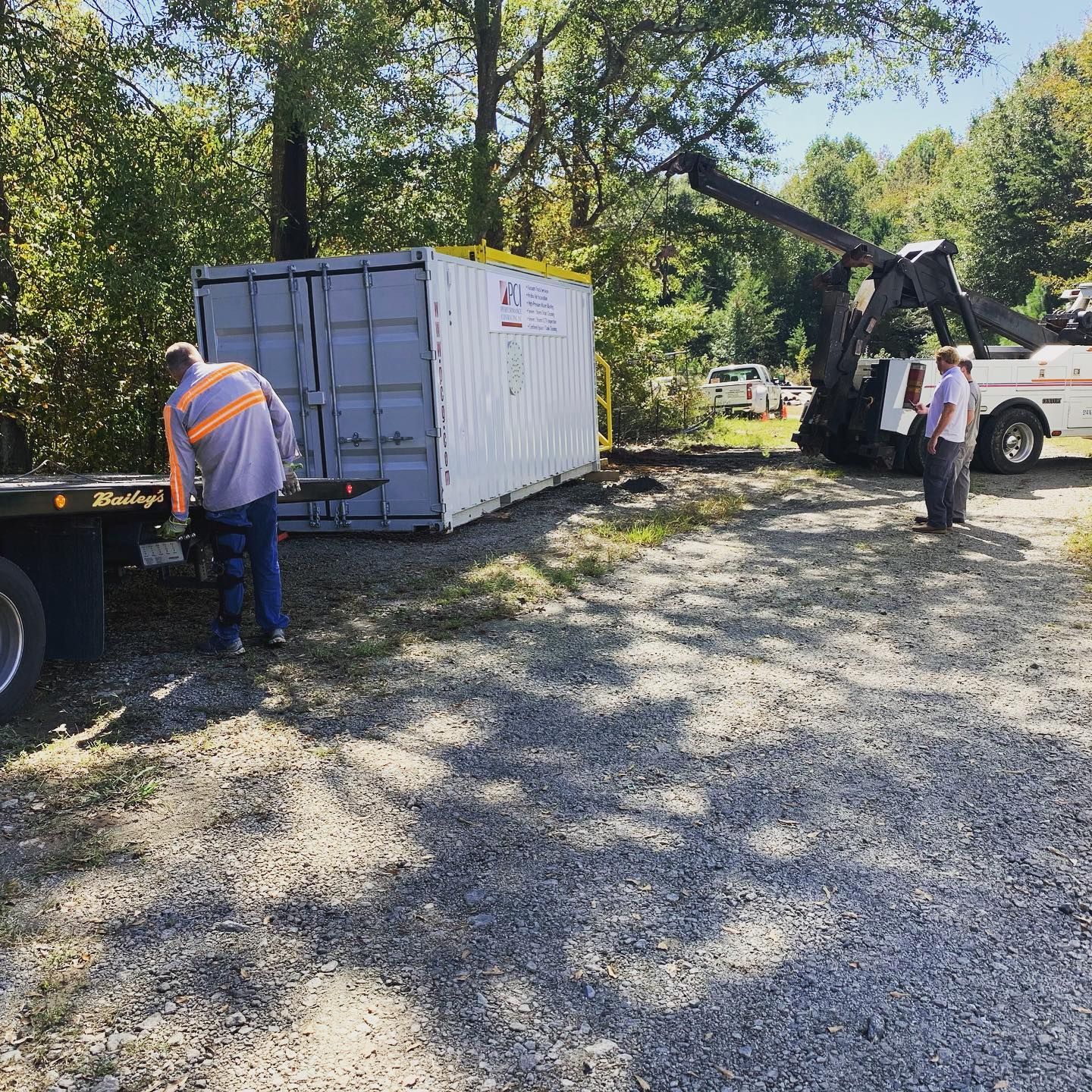 A tow truck lifting a shipping container onto a flatbed truck in a gravel lot, with two men observing and a third working.
