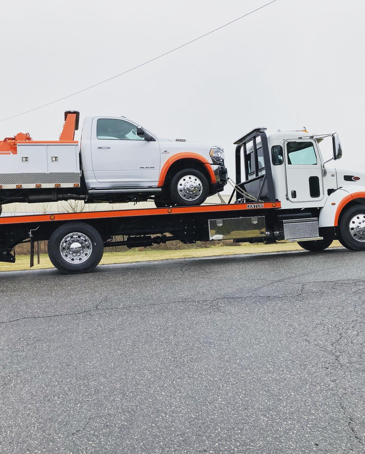 White tow truck carrying a white pickup truck on a flatbed. The setting is a paved road under a cloudy sky.