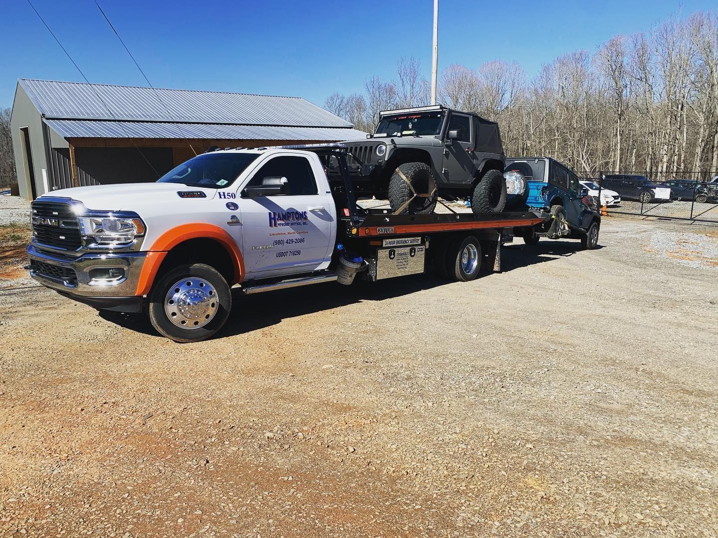 A tow truck carries a black Jeep and a van on a flatbed. The truck is white and orange, parked outside on a gravel lot.