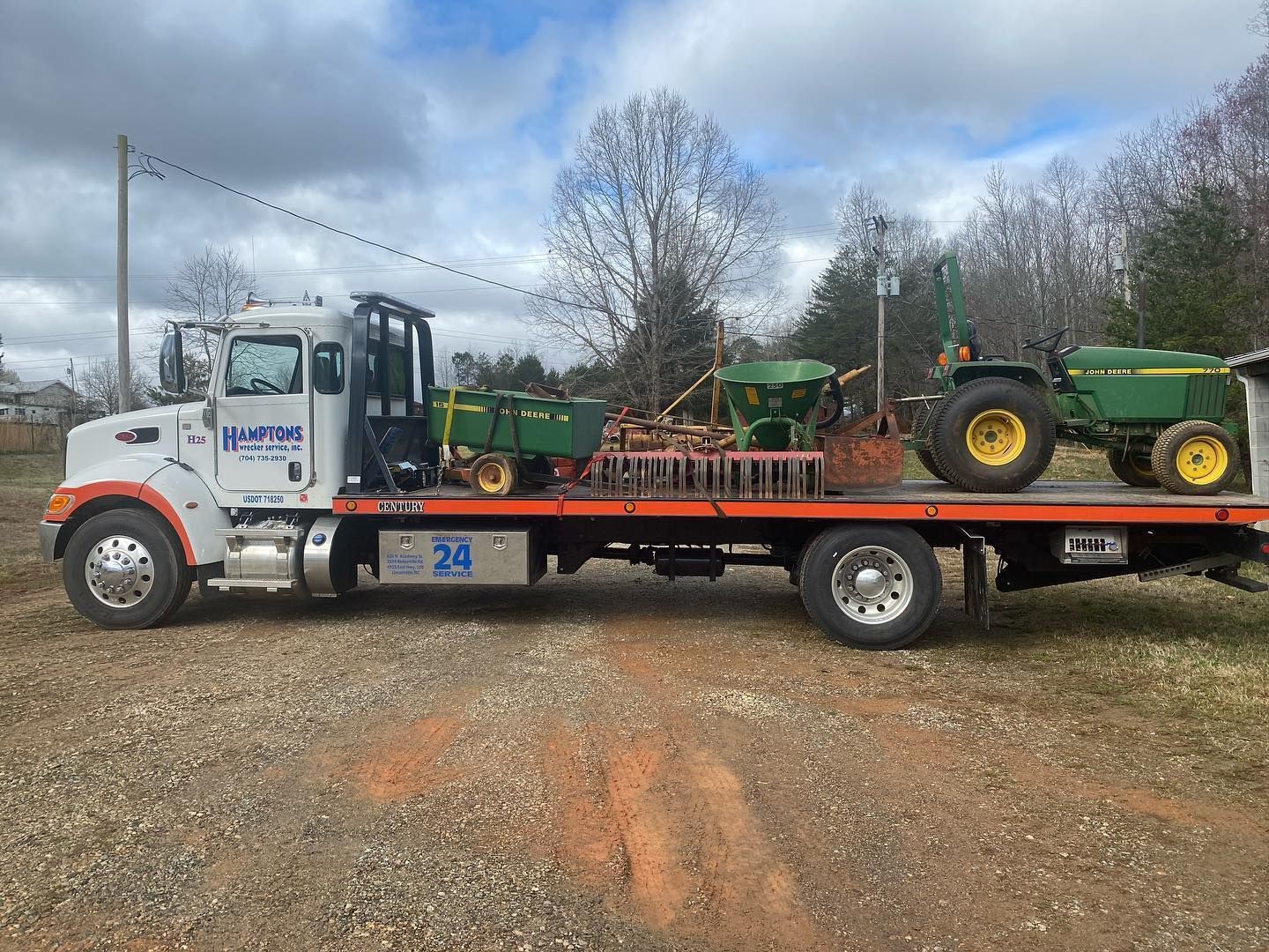 A white tow truck transports farm equipment on a red flatbed. The scene is outdoors, with a cloudy sky.