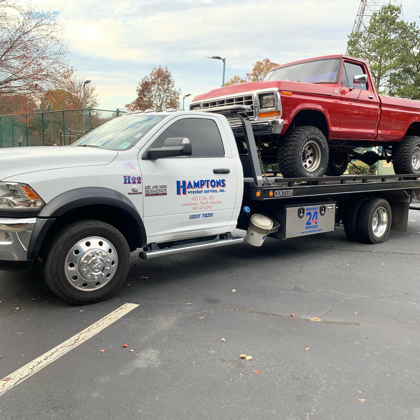White tow truck hauling a red vintage pickup truck. Hamptons Towing logo visible on the truck's door, parked on asphalt.
