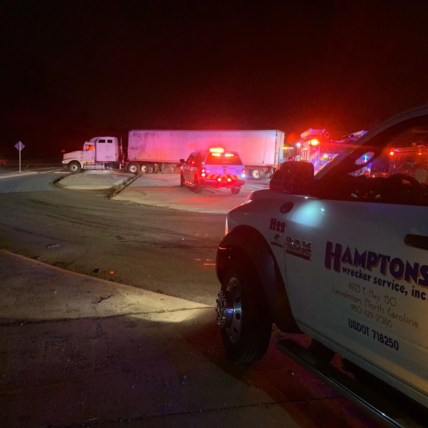 Night scene of a semi-truck and emergency vehicles at an accident site. A tow truck is in the foreground.