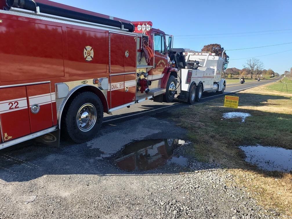 Red fire truck towing a white tow truck on a gravel shoulder next to a road on a sunny day.