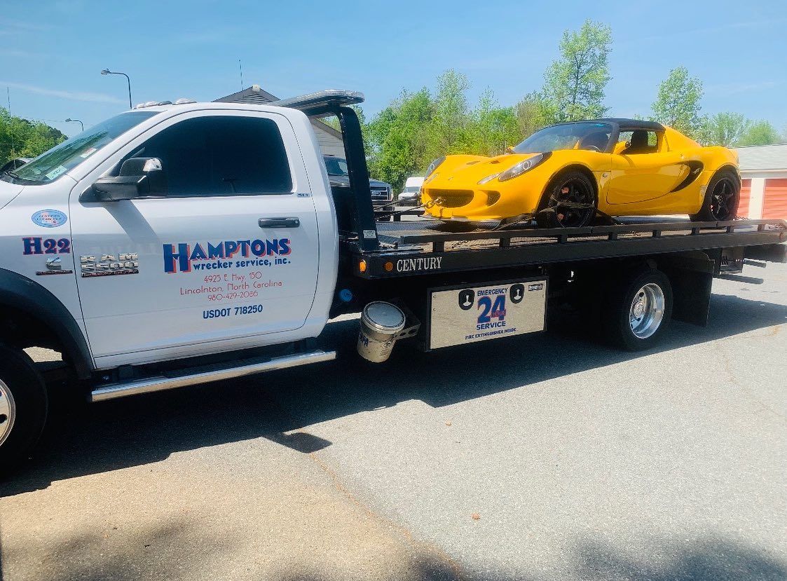 A yellow Lotus sports car being towed by a white Hamptons Towing truck on a gravel surface under a blue sky.