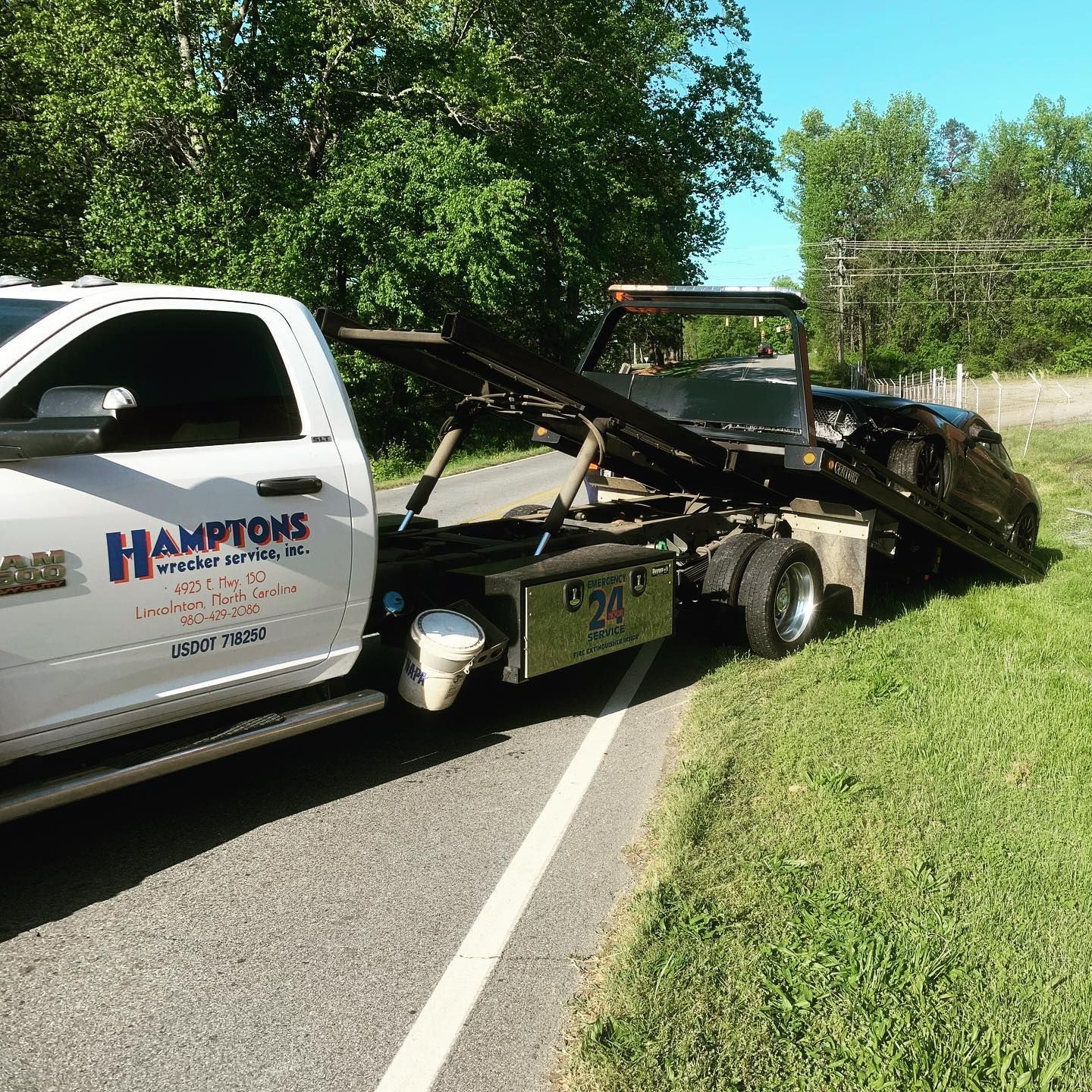 White tow truck from Hamptons Wrecker Service towing a dark-colored vehicle off a grassy shoulder of a road.