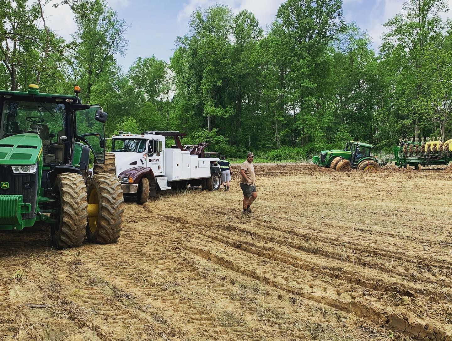 A tractor pulls a white truck from a muddy field, with another tractor planting crops in the background. 