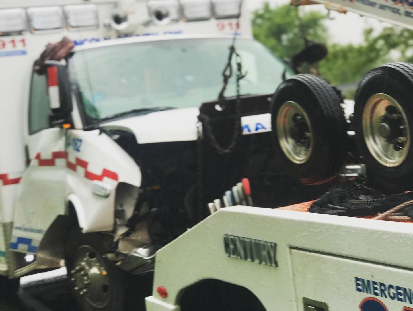 Damaged ambulance being towed away after an accident; white, red, and black colors visible; tow truck in foreground.