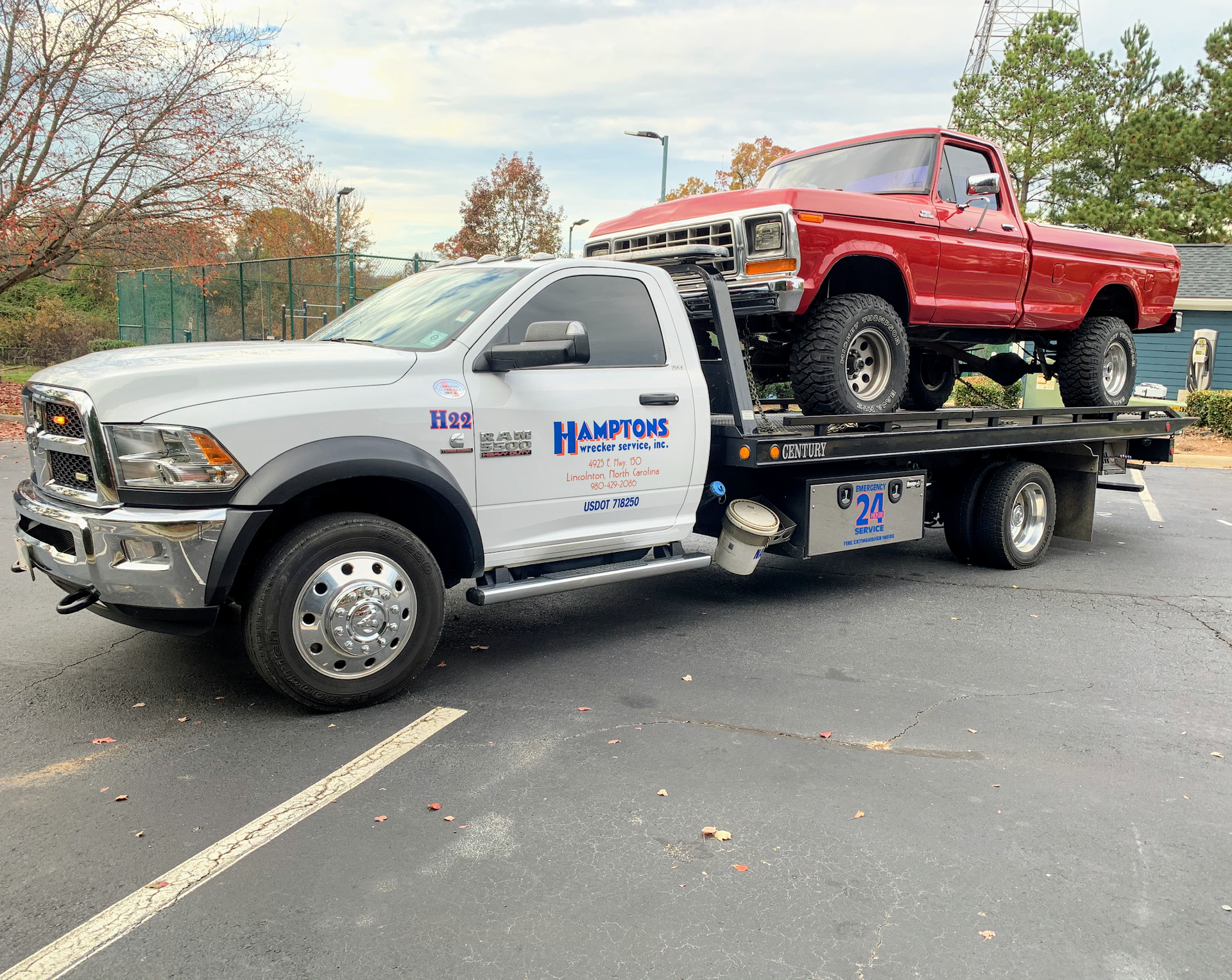 A white tow truck transports a red pickup truck on its flatbed in a parking lot. The setting is outdoors with fall foliage.
