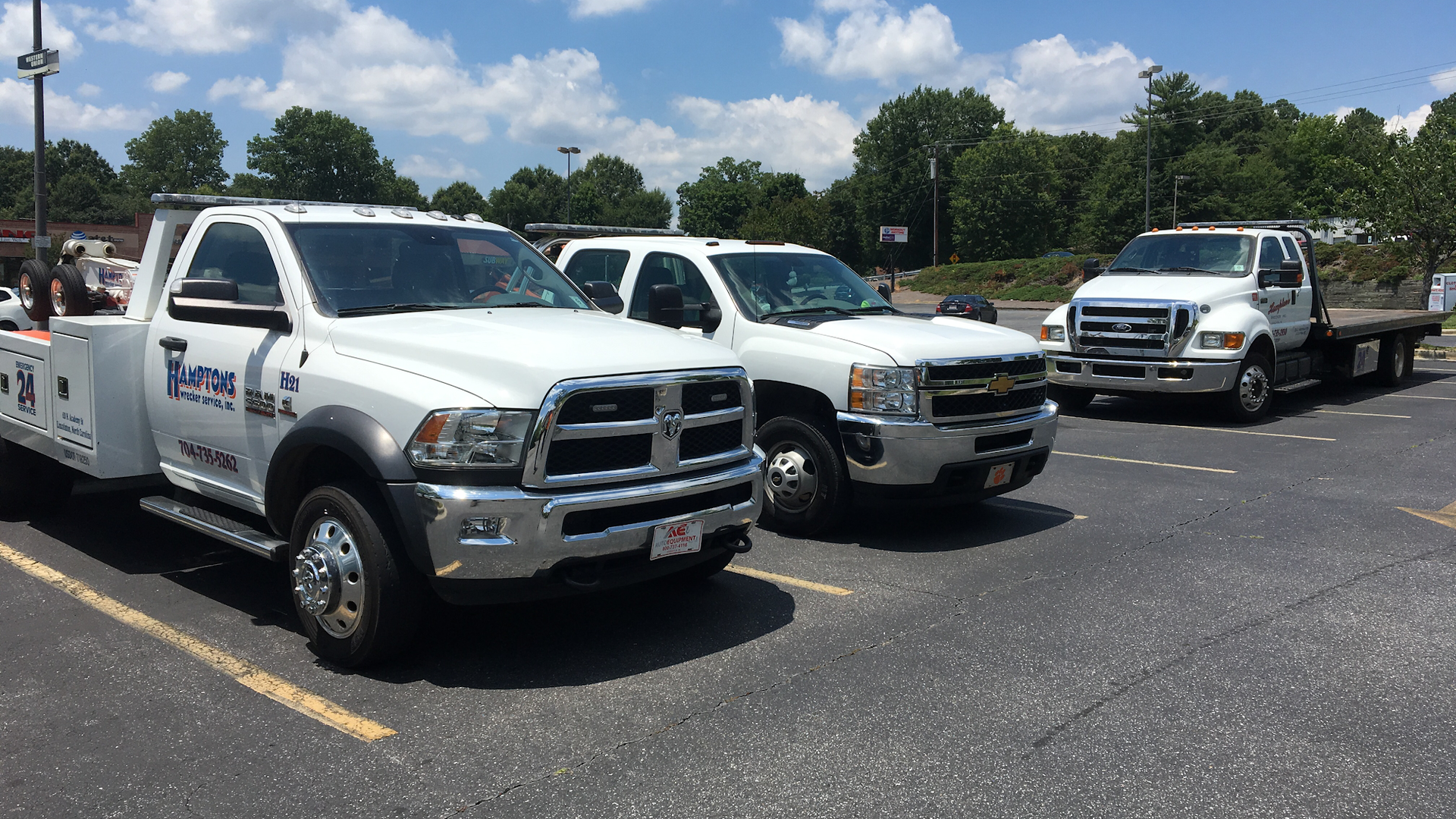 Three white tow trucks parked in a lot on a sunny day. Trees and a building are visible in the background.