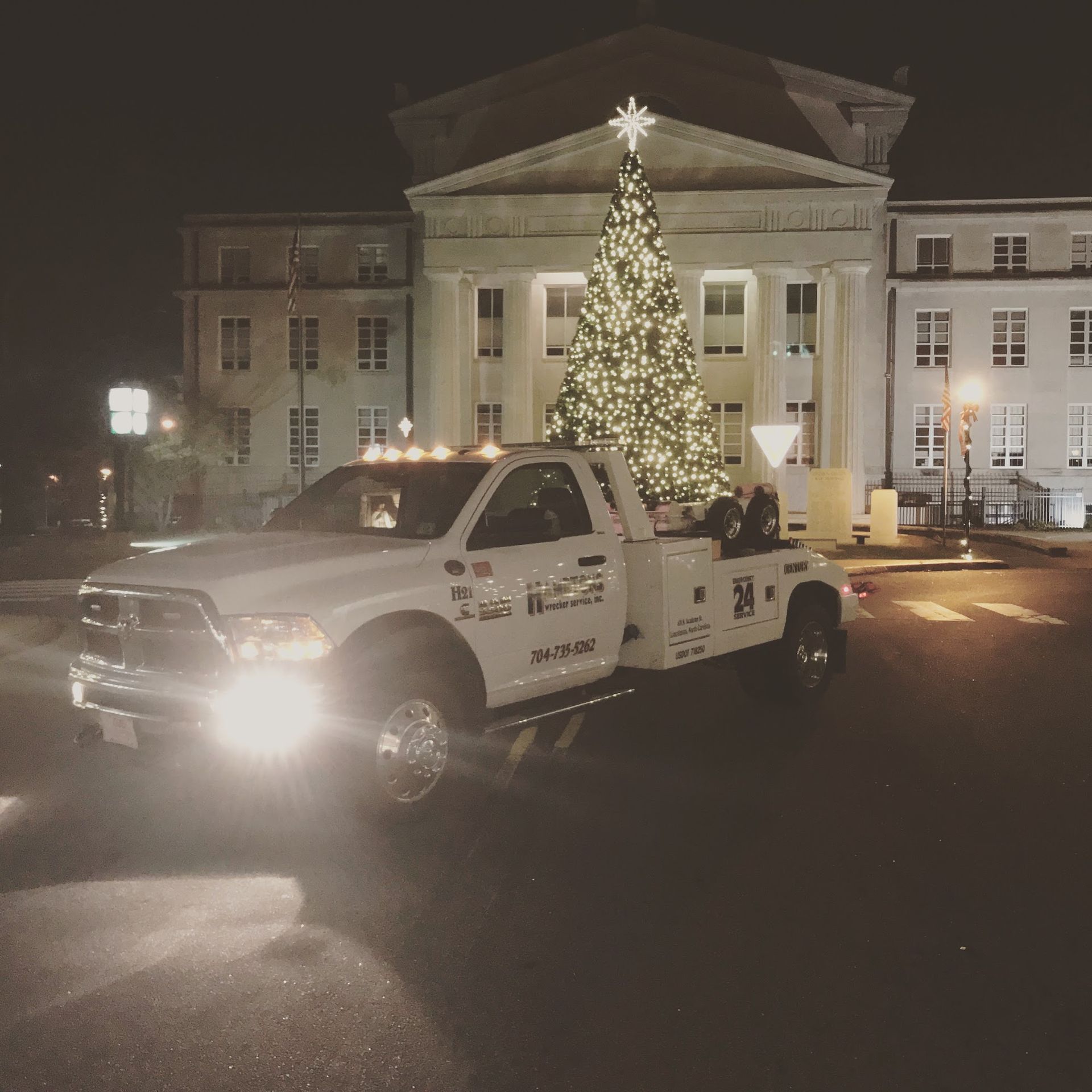 A white tow truck parked in front of a building with a lit Christmas tree at night.