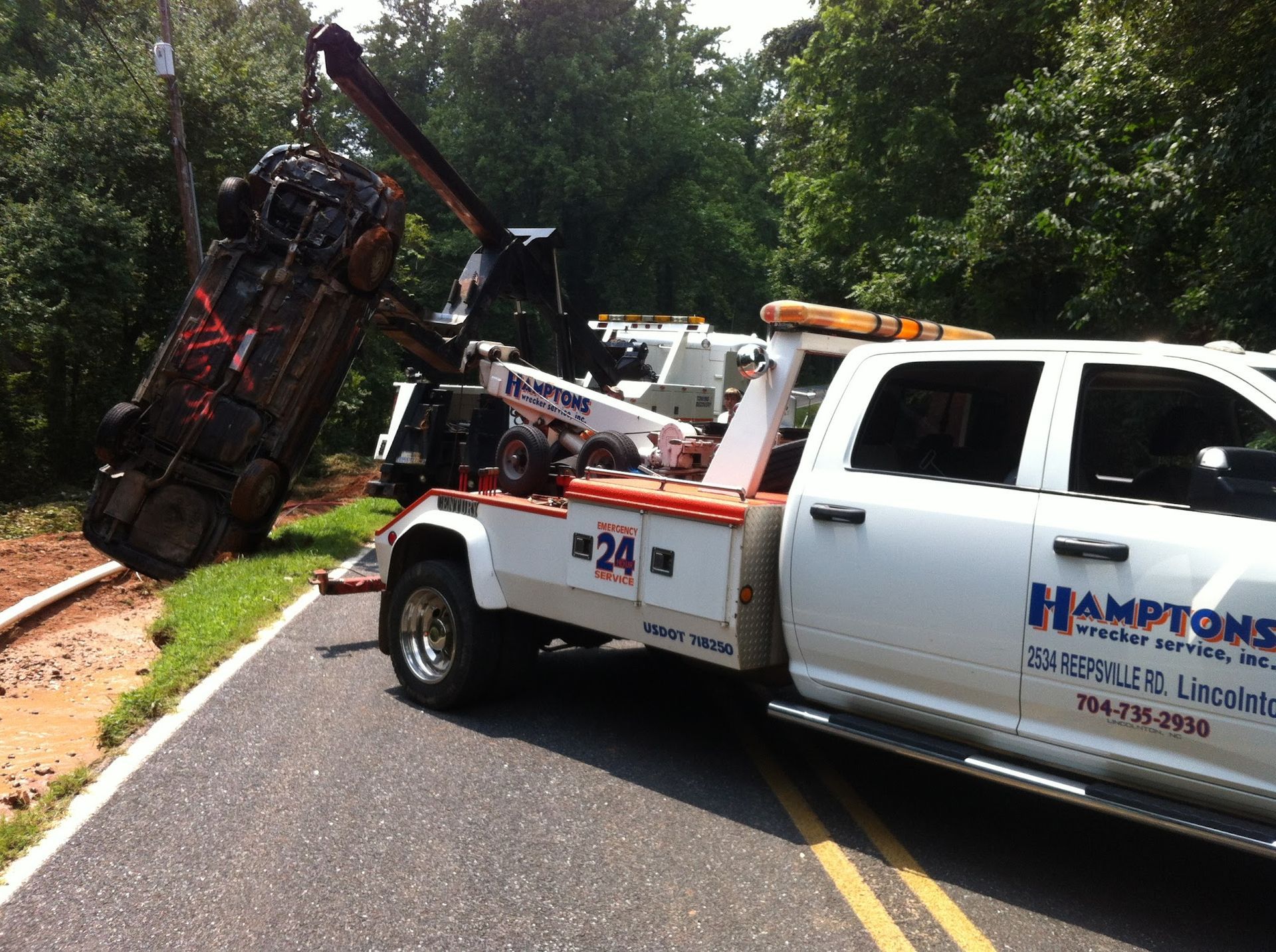 A tow truck lifts a damaged vehicle from a roadside. The white truck has a red and orange stripe.