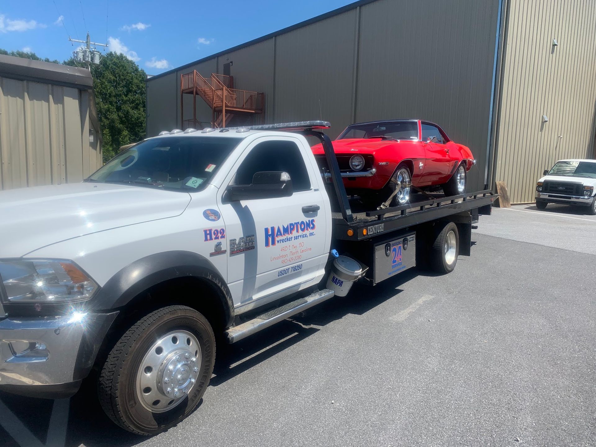 White tow truck hauling a red classic car in a parking lot. The truck has the company name 