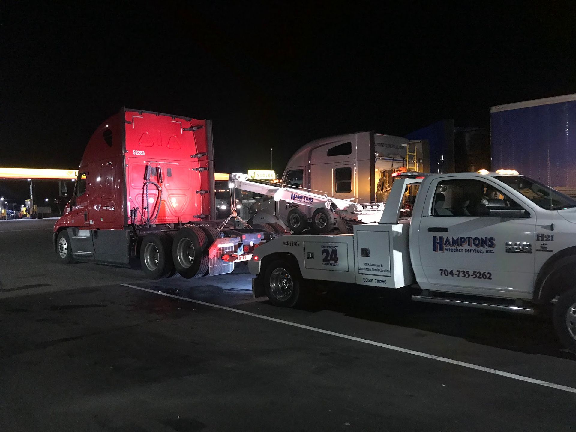 A red semi-truck being towed by a white tow truck at night. The tow truck has the business name 