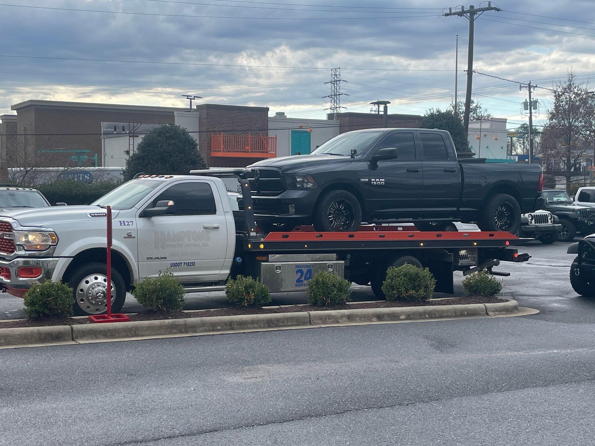 A black pickup truck being towed on a flatbed tow truck. The tow truck is white and parked in a lot on an overcast day.