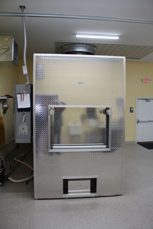 A large, stainless steel freezer with a window and door in a room. A ventilation pipe is above the freezer.