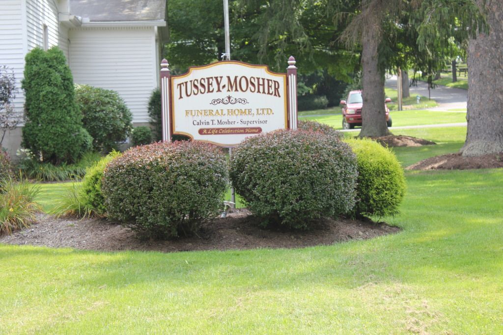 Sign for Tussey-Mosher Funeral Home with green bushes and lawn in front. A red car is visible in the background on a road.