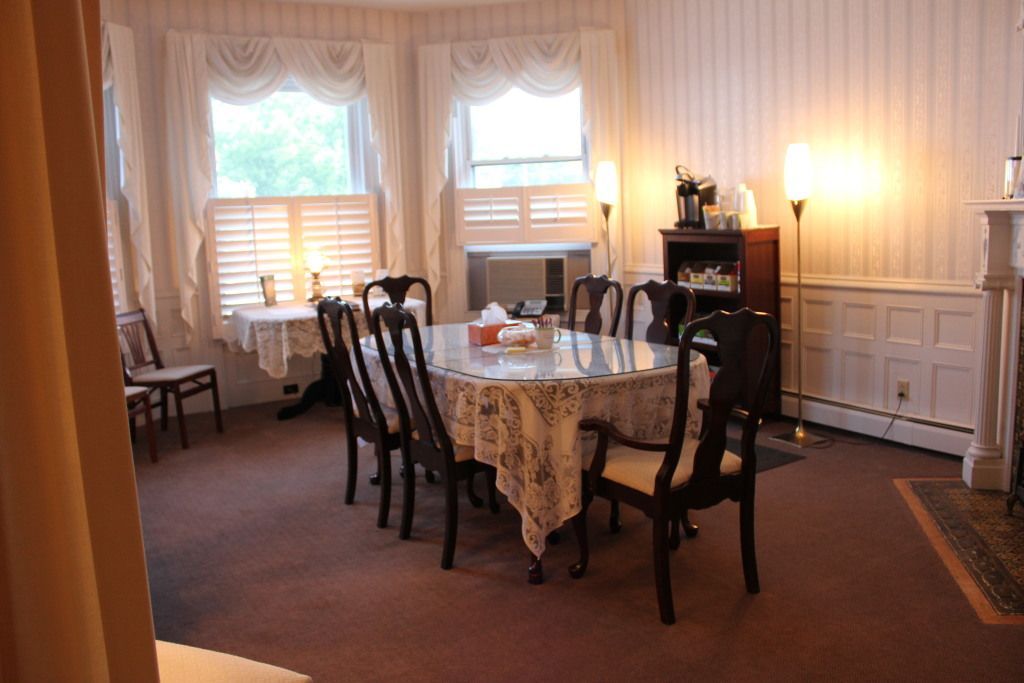 Dining room with a large oval table set for a meal. Windows with lace curtains, dark wood chairs, and a fireplace.