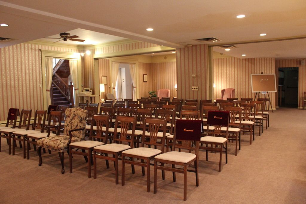 A room with rows of chairs, likely prepared for a formal event. Beige wallpaper and carpeting, warm lighting.