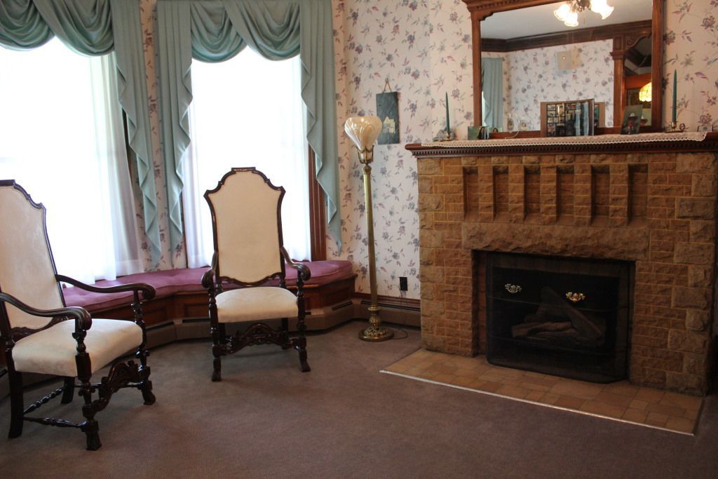 A Victorian-era living room with a stone fireplace, two ornate chairs, and large windows with blue drapes. The room has floral wallpaper.