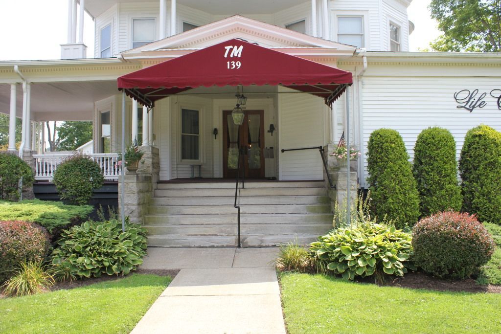Exterior view of a white building with a burgundy awning over the entrance, a set of stairs, and landscaping. 