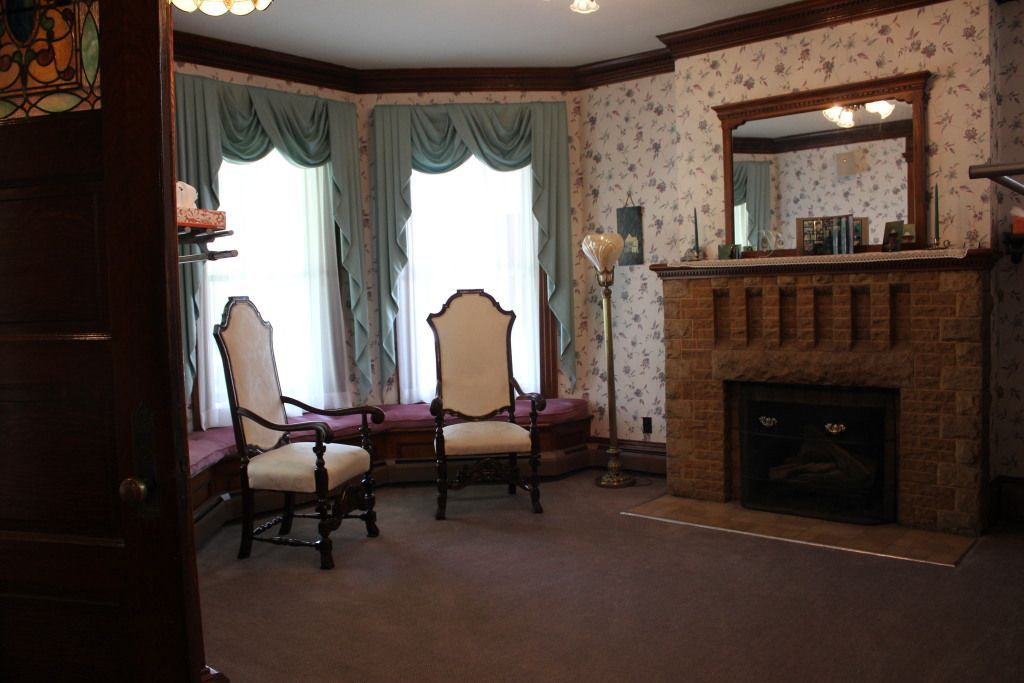 A Victorian-style room with a bay window, fireplace, two chairs, and floral wallpaper.