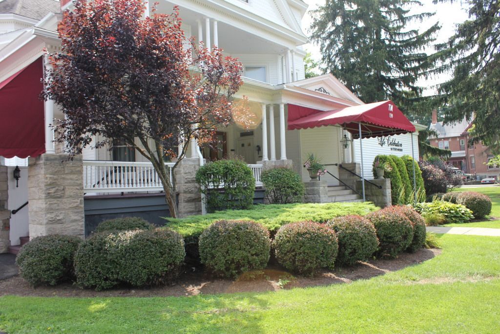 A white Victorian-style house with red awnings. Lush green lawn and shrubs frame the porch with white columns and a dark red-leaved tree.