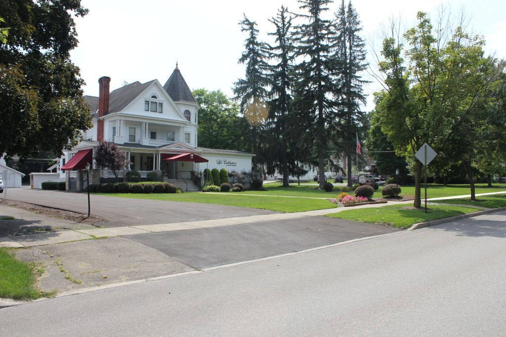 White Victorian house with red awnings, lawn, trees, and street in a residential setting.