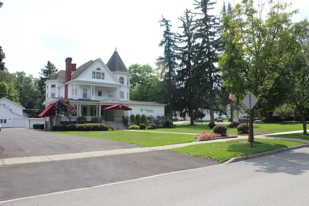 White Victorian house with a tower, red awning, and a well-manicured lawn; trees and a street visible in the foreground.
