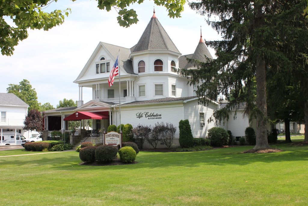 White Victorian building with turrets and a red awning, American flag on the porch, set on a green lawn.