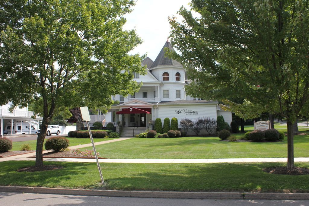 White Victorian-style building with a red awning and a small tower, nestled behind green trees and a well-maintained lawn.