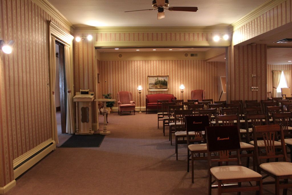 A funeral home chapel with rows of chairs, a red sofa, and floral wallpaper. Soft lighting and neutral tones create a somber atmosphere.