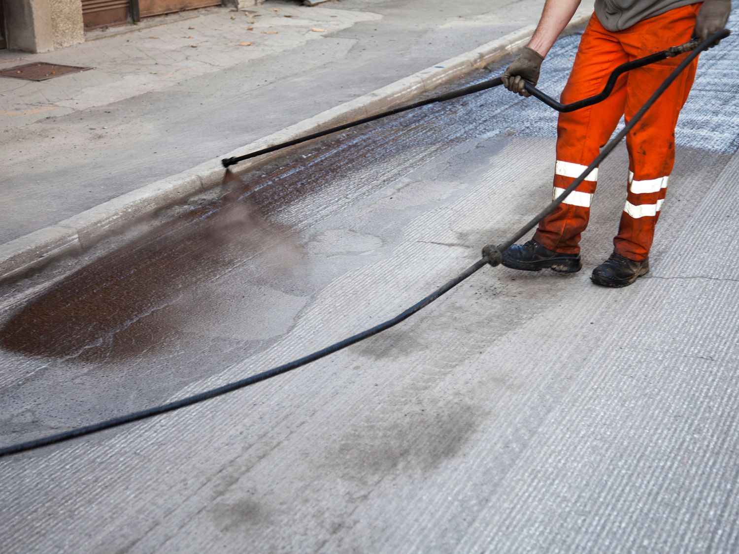 A man in orange pants is spraying asphalt on the ground with a hose.