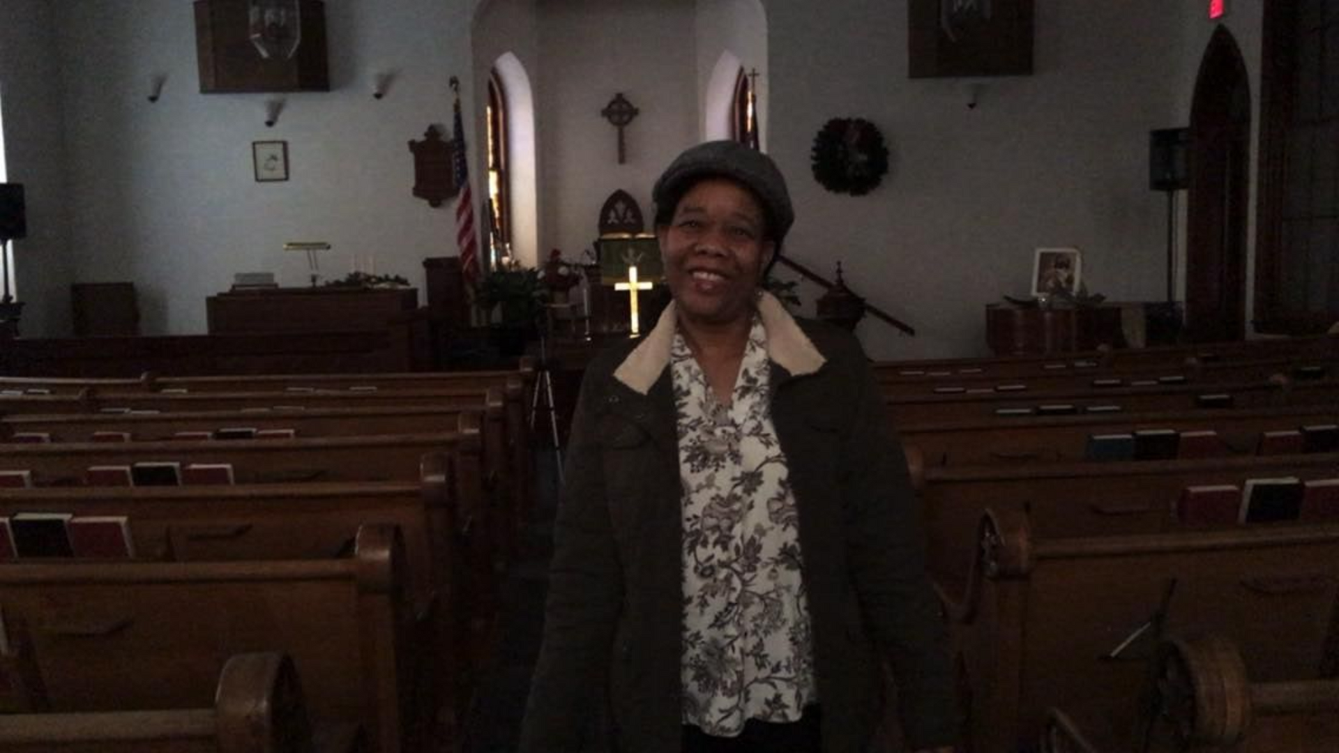 Woman smiling in church, wearing hat and coat, wooden pews in the background.