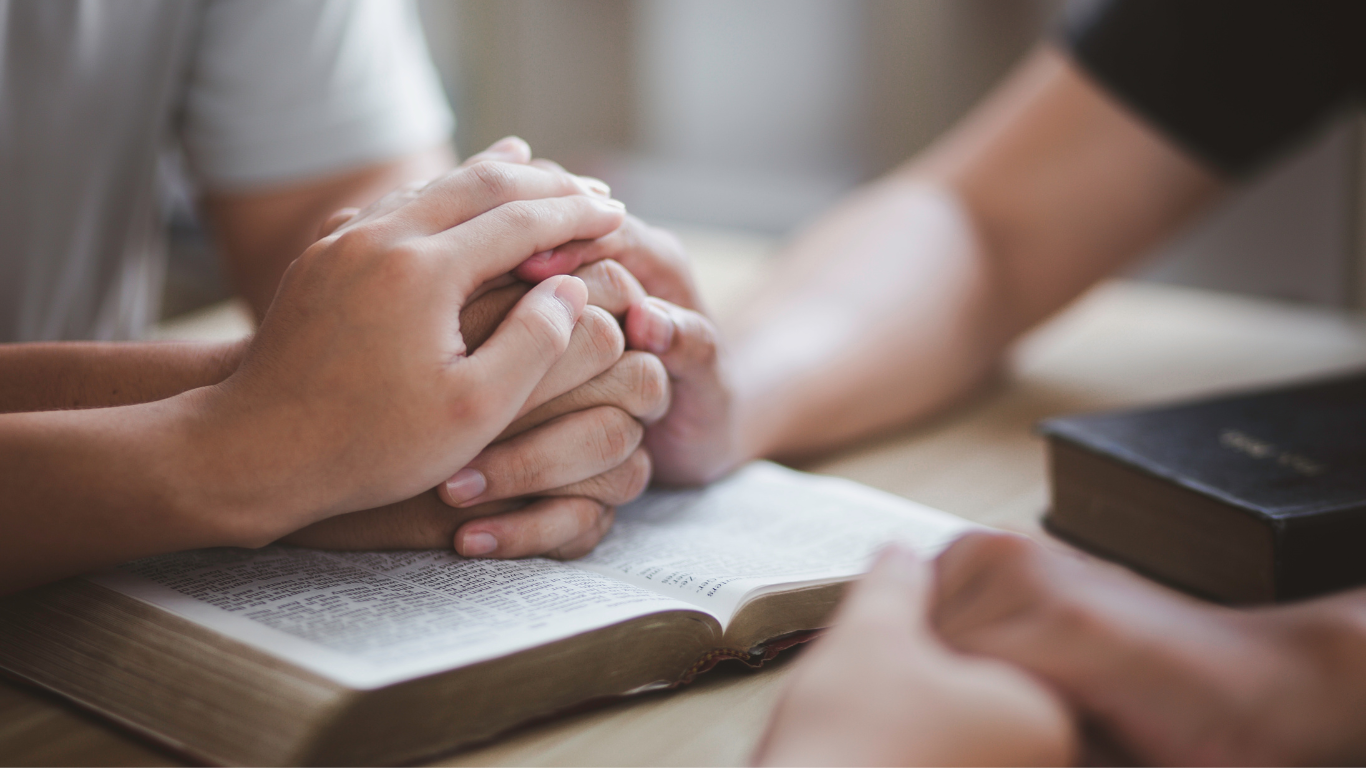 Hands clasped in prayer over an open book, with a Bible on a wooden surface.