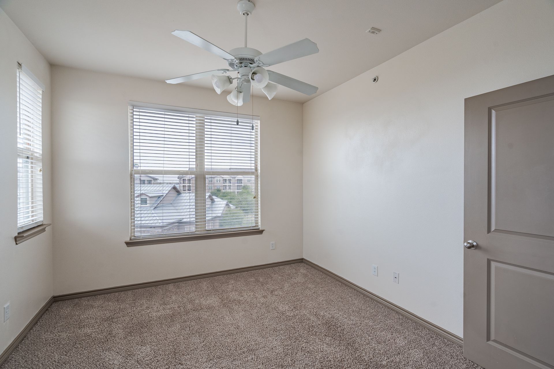 Empty carpeted bedroom with two windows, blinds, and a ceiling fan.
