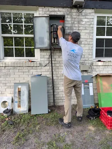Electrician working on an electrical panel on a brick wall outside. He wears a gray shirt and khaki pants.