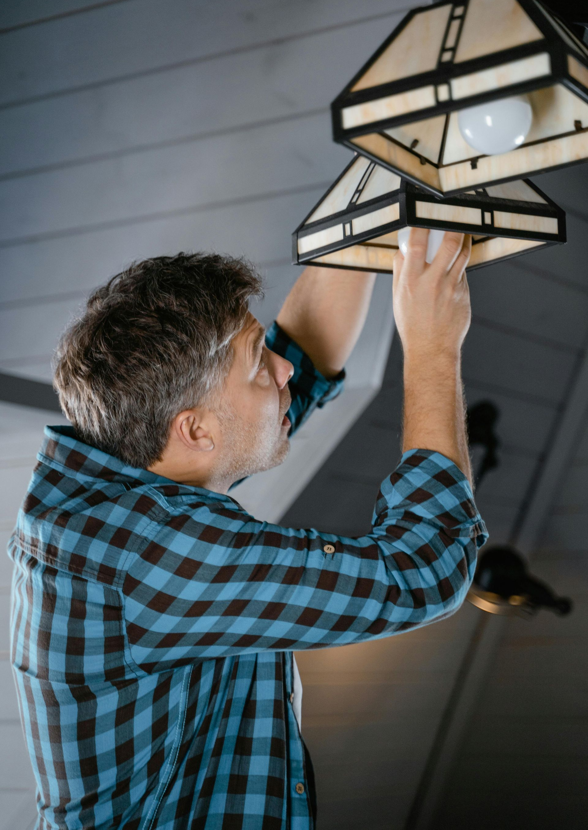 Man in blue plaid shirt changing a lightbulb in a wooden fixture indoors.