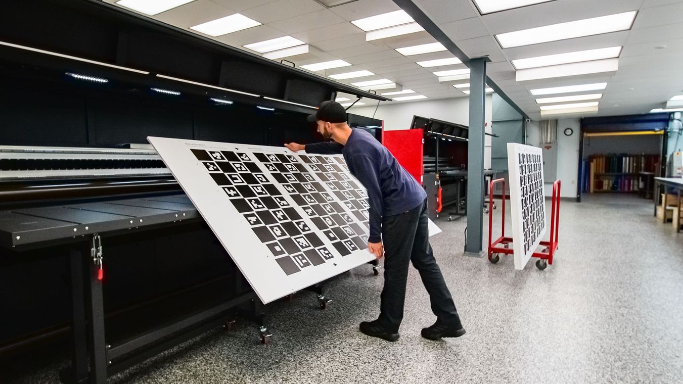Man in a workshop holding a large white panel with black patterns, possibly for calibration, by a black machine.