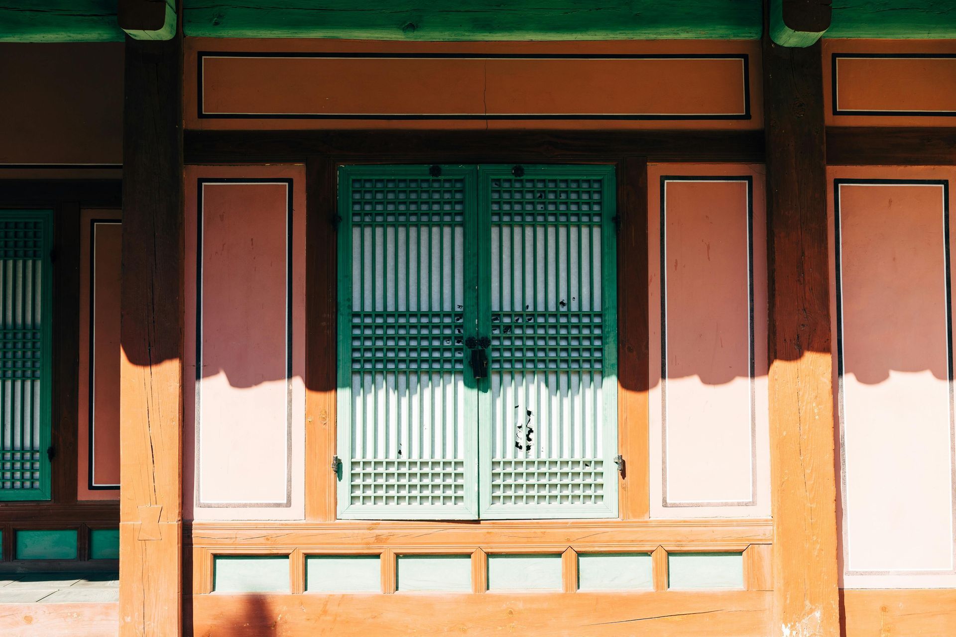 Traditional Korean building exterior. Teal doors and window, pink walls, orange and brown trim, sunlight.