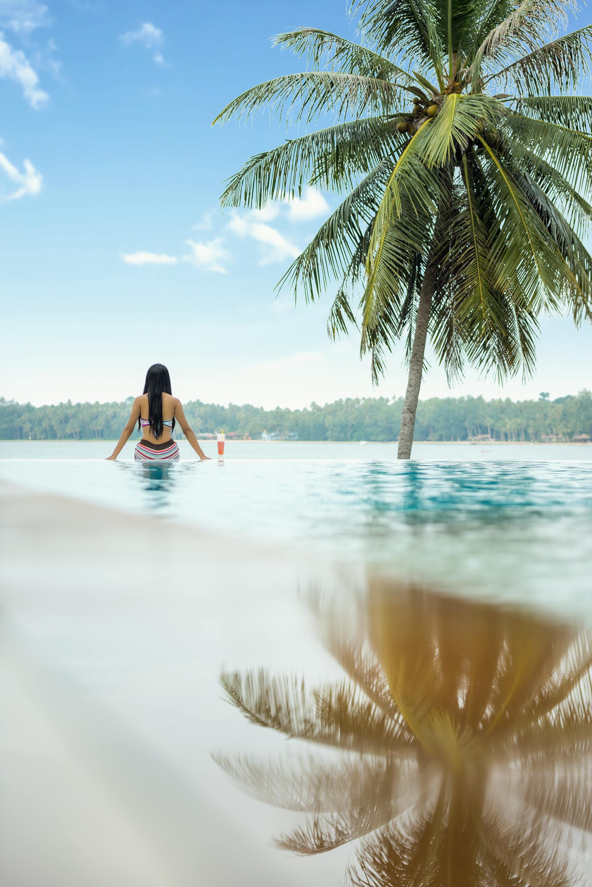 Una mujer está sentada en una piscina junto a una palmera.