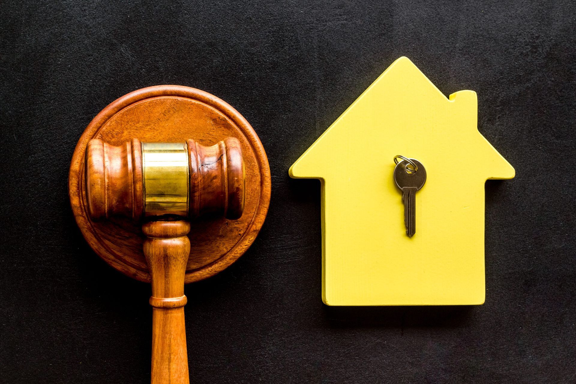 Wooden gavel next to a yellow house-shaped cutout with a key, on black background.