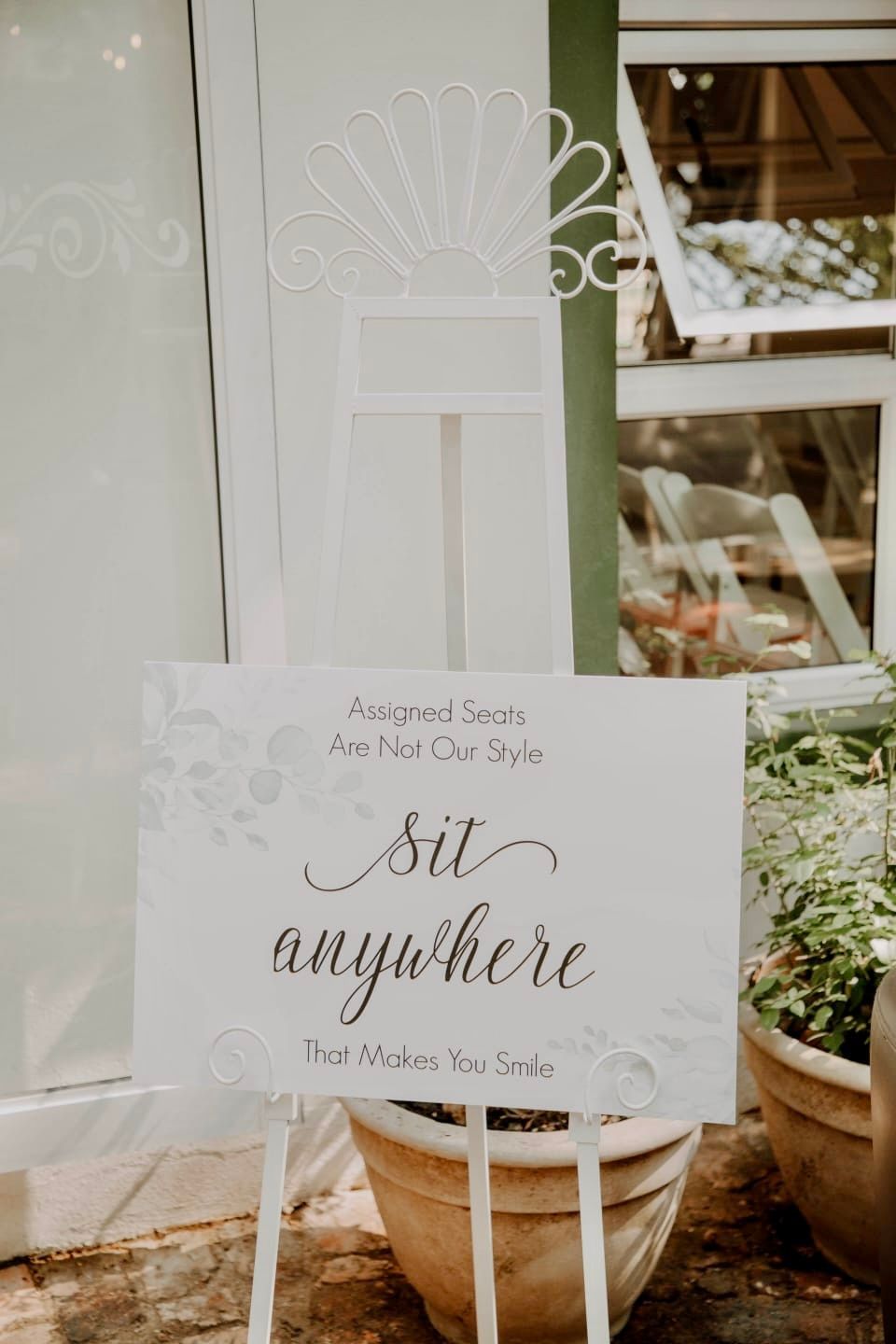 A sign that says `` sit anywhere '' is sitting on a white easel in front of a potted plant.