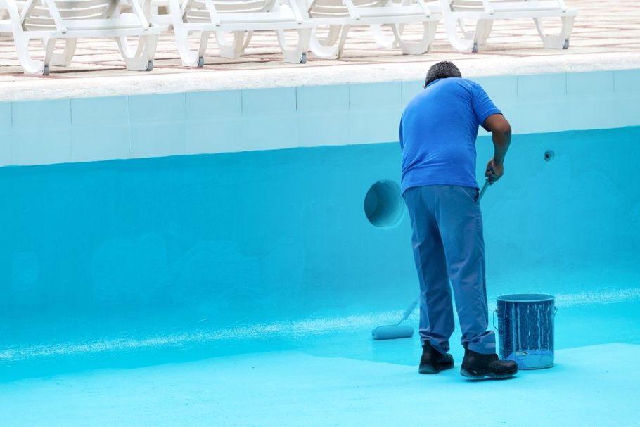 Worker cleaning the side of an empty blue swimming pool with a bucket nearby