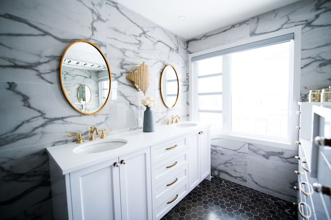 black tiled bathroom with a white sink and countertop, gold framed mirrors with window with sunlight
