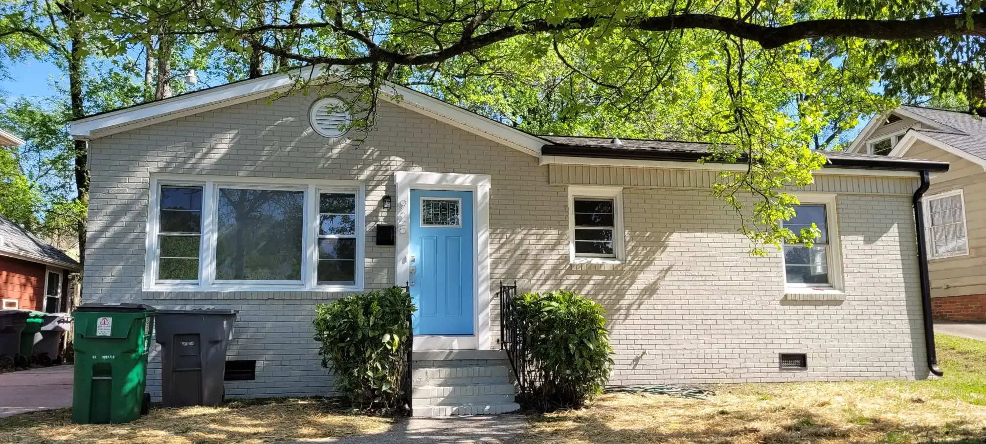 A white house with a blue door and a green trash can in front of it.