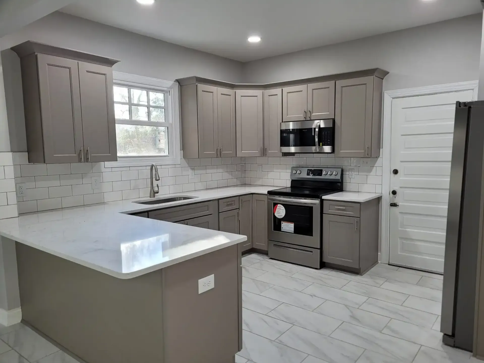 A kitchen with gray cabinets , white counter tops , stainless steel appliances and a large island.