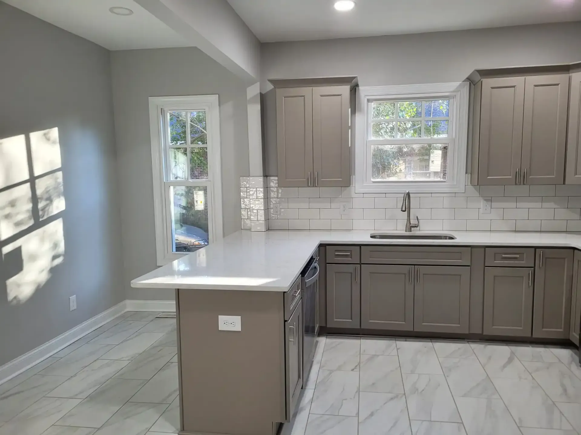 A kitchen with gray cabinets , white counter tops , a sink and a window.