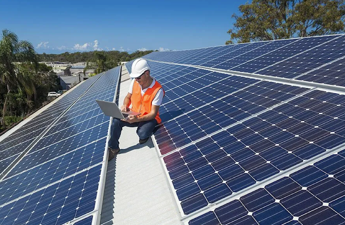 A man is sitting on top of a roof with solar panels and using a laptop.