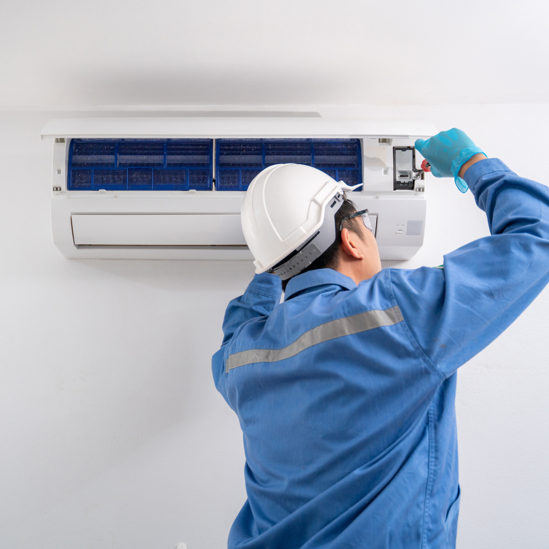 HVAC technician in blue coveralls, gloves, and hard hat working on a wall-mounted air conditioner.